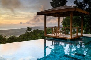 A wooden gazebo with two wicker chairs overlooking a clear blue infinity pool.