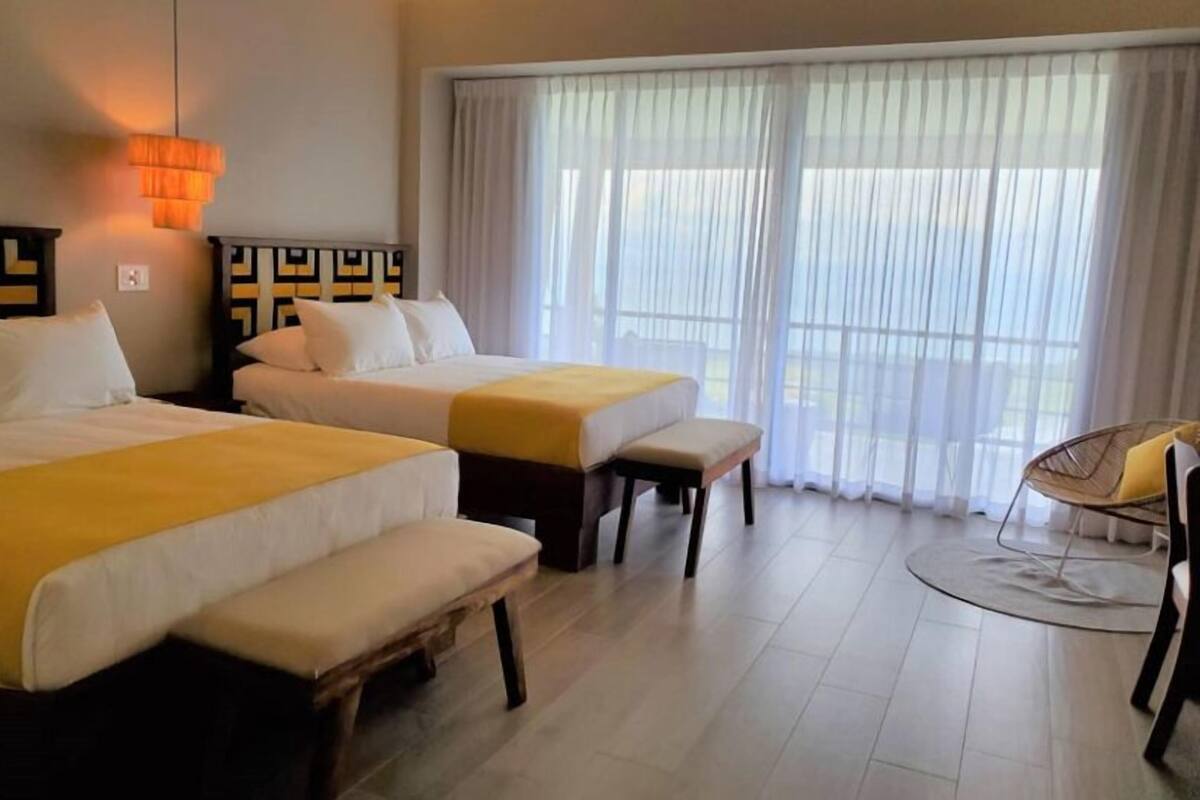 A hotel room with two beds, a bench, a chair, and a large window with curtains.