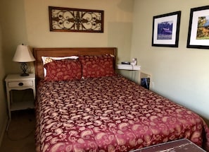 A neatly made bed with a patterned bedspread, a bedside lamp, a small shelf with a decorative item, and two framed pictures on the wall.