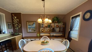 Une salle à manger avec une table en bois, des chaises blanches, un lustre et une bibliothèque contenant divers objets.