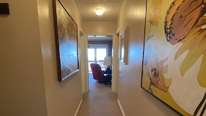 Un couloir doté d’un sol recouvert de moquette, des œuvres d’art encadrées accrochées aux murs, et une vue sur une pièce où l’on aperçoit une chaise et une lampe.
