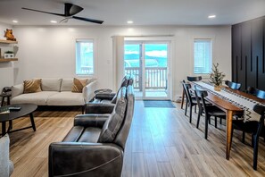 A modern living room with a sofa, a dining table with chairs, a ceiling fan, and a view through a glass door to the outside.