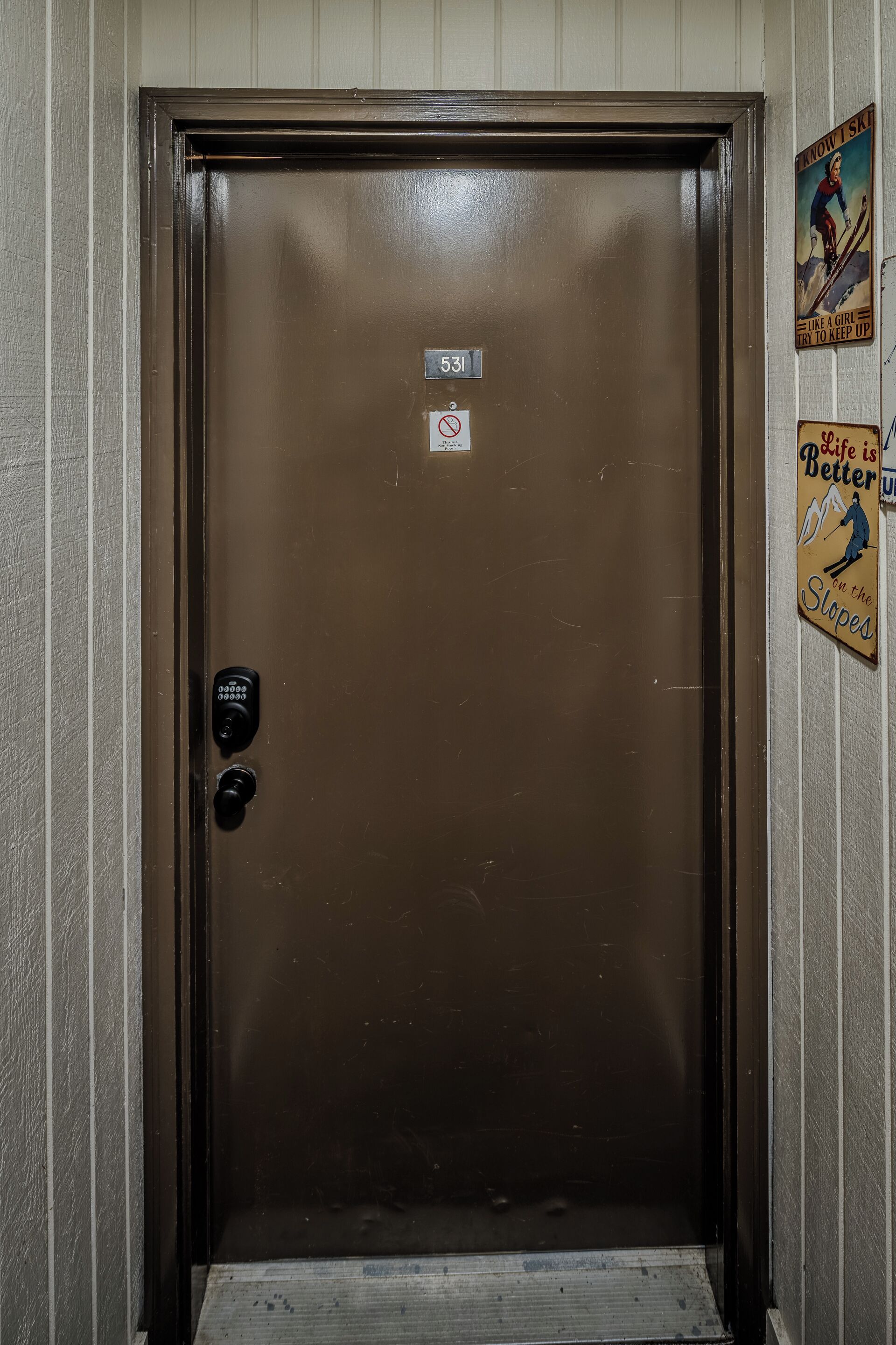 Una puerta de ascensor marrón con un panel numérico de bloqueo, un cartel con el número 531, y carteles temáticos de esquí de estilo antiguo en la pared.