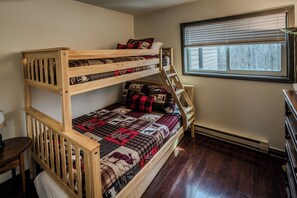 A bunk bed with a quilt, a window with blinds, and a wooden desk.
