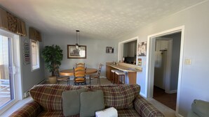 A living room with a plaid sofa, a dining table with chairs, a kitchen area, and a hallway. - Sugar Ski Comfort Two Bedroom - Unit 631 (Sugar Mountain)