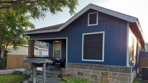 Een blauw huis met een overdekte veranda, een fundering van baksteen en een kleine tuin met een palmboom.