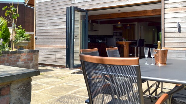 Une terrasse extérieure avec une table et des chaises, un mur en pierre et un bâtiment en bois doté d’une porte coulissante.