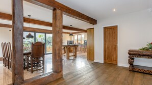 Une pièce spacieuse avec des poutres apparentes, un coin repas avec des chaises, une cuisine à l’arrière-plan et une porte en bois.
