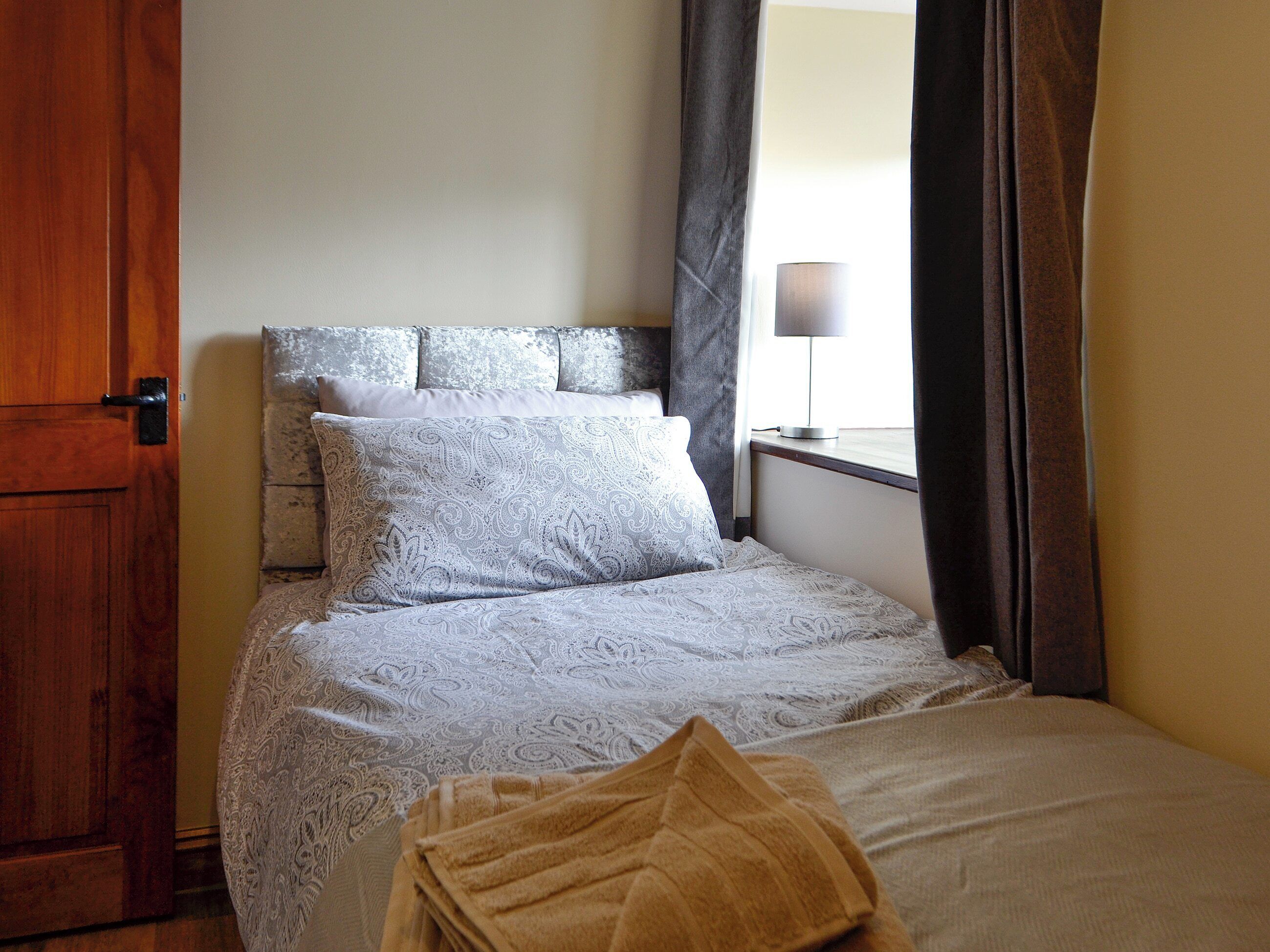 Een slaapkamer met een bed, een houten deur, een nachtkastje met een lamp en een raam met gordijnen.