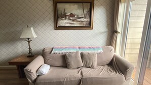 Un salon avec une table basse en bois, un canapé beige agrémenté de coussins, un tableau encadré au-dessus du canapé, une table d’appoint en bois avec une lampe, et une fenêtre avec des rideaux.