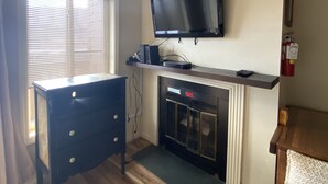 Une pièce avec une cheminée, un téléviseur fixé au mur, une commode en bois et une fenêtre avec des stores.