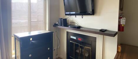 Una habitación con chimenea, televisor montado, cómoda de madera y ventana con persianas.