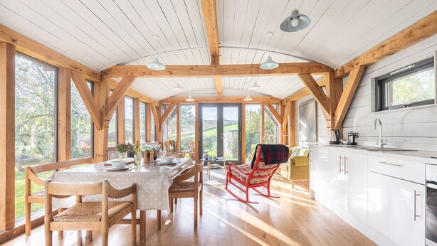 Una cocina amplia con vigas de madera, un comedor con una mesa dispuesta para cuatro personas y ventanas grandes que ofrecen vistas al exterior.
