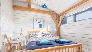 Un dormitorio con una cama de madera, un edredón a cuadros y una colcha azul. Se ve una ventana con cortinas estampadas, un escritorio blanco y una silla.