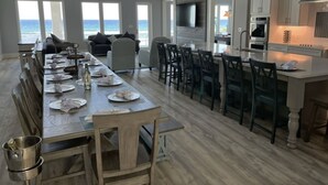 A spacious dining area with a long table set for a meal, a kitchen with modern appliances, and large windows offering ocean views.