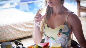 Una persona sentada en una mesa con diversos alimentos y un vaso de bebida, con una piscina y vegetación de fondo.