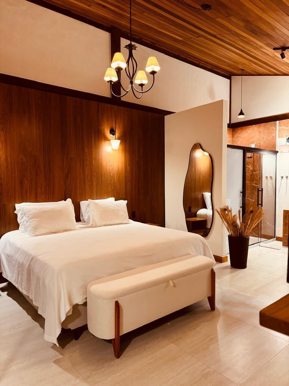 A modern bedroom with a large bed, a wooden ceiling, a wall-mounted light fixture, a mirror, and a bathroom visible through a glass door.