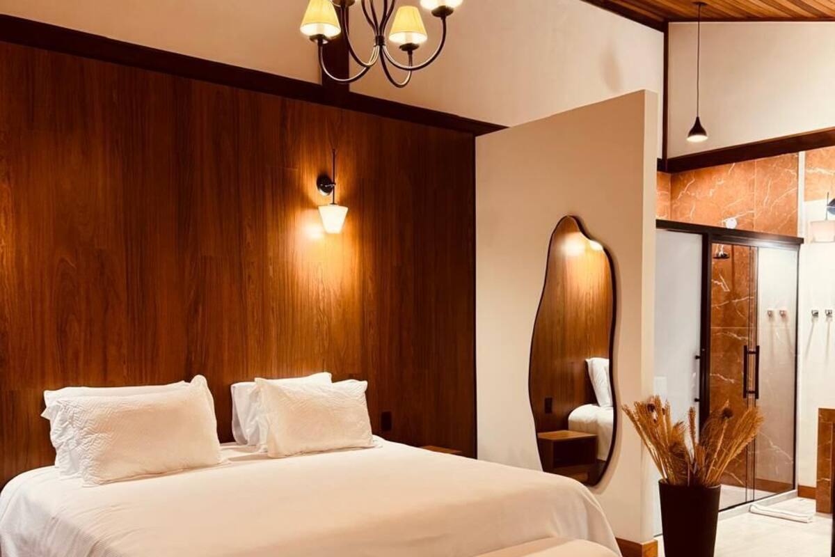 Ein modernes Schlafzimmer mit einem großen Bett, einer Holzbalkendecke, einer Wandleuchte, einem Spiegel und einem durch eine Glastür einsehbaren Badezimmer.