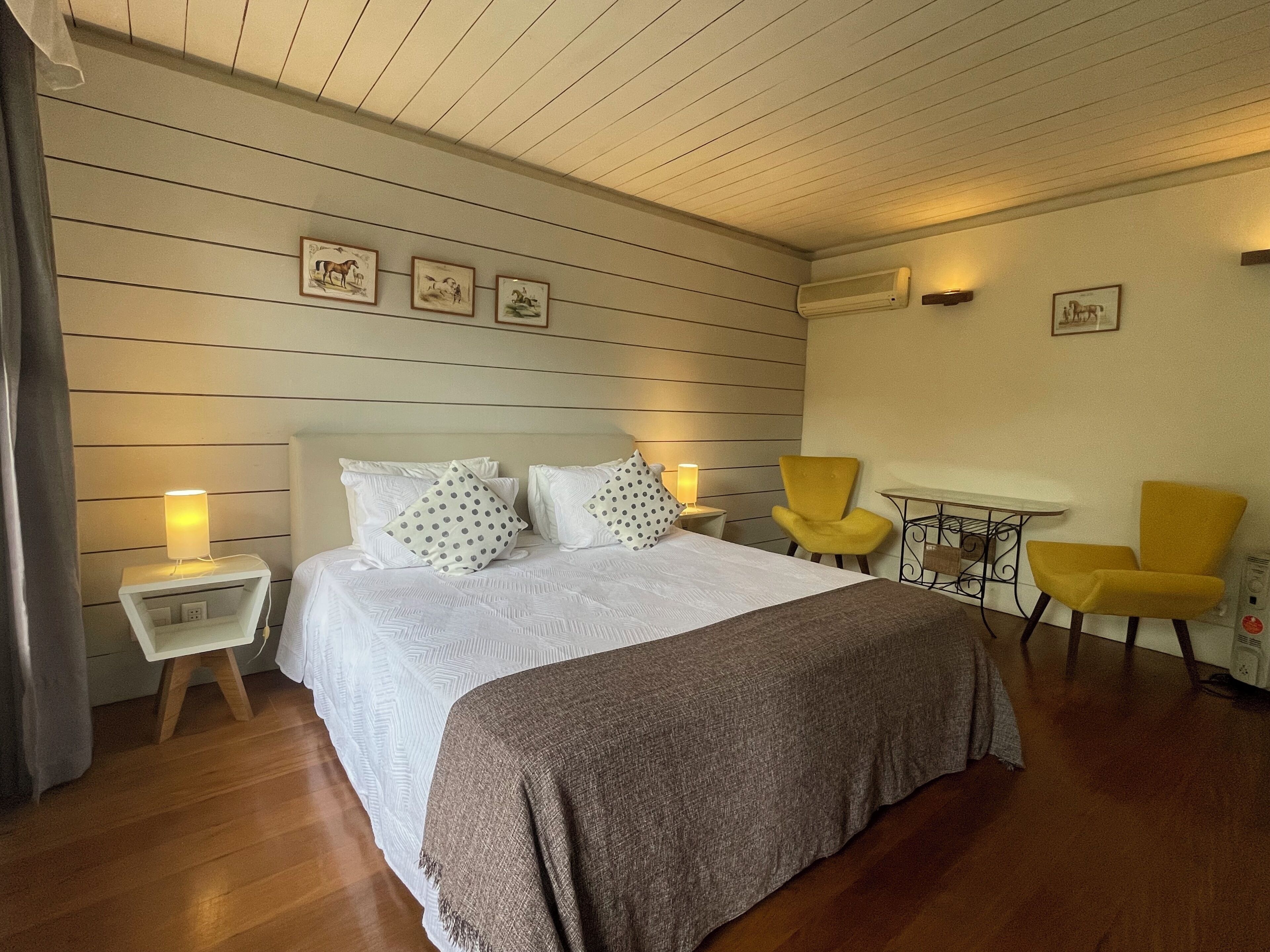 A hotel room with a bed, two bedside tables with lamps, a small table with a chair, and a wall-mounted air conditioner.