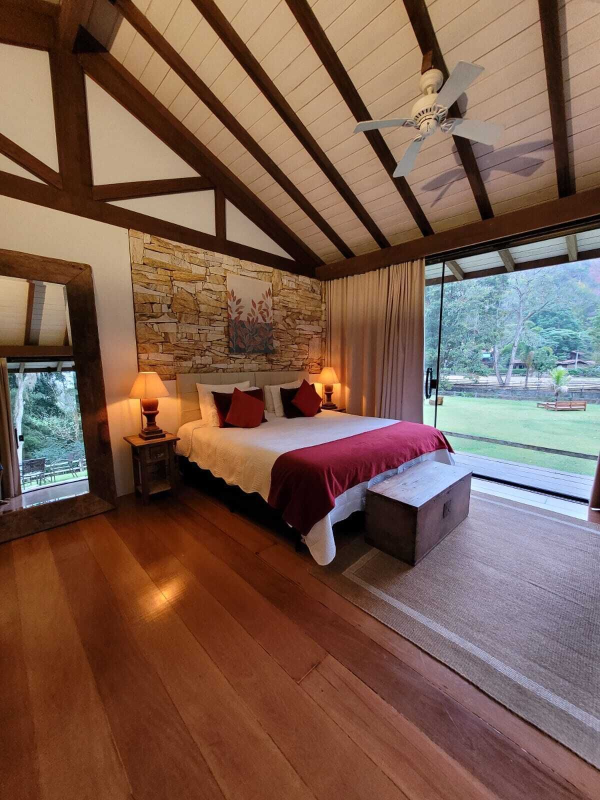Ein Schlafzimmer mit Steinwand, Holzbalken, einem Bett mit roter Bettwäsche, einem Nachttisch, einer Lampe und Blick ins Freie.