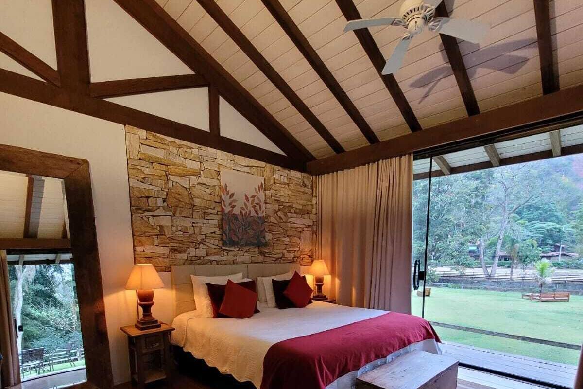 Ein Schlafzimmer mit Steinwand, Holzbalken, einem Bett mit roter Bettwäsche, einem Nachttisch, einer Lampe und Blick ins Freie.