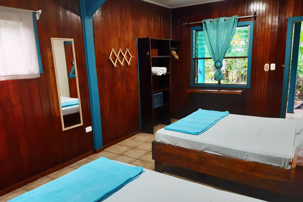 Una habitación con paredes de madera, una cama con una colcha azul, un armario, un espejo y una ventana con cortinas.