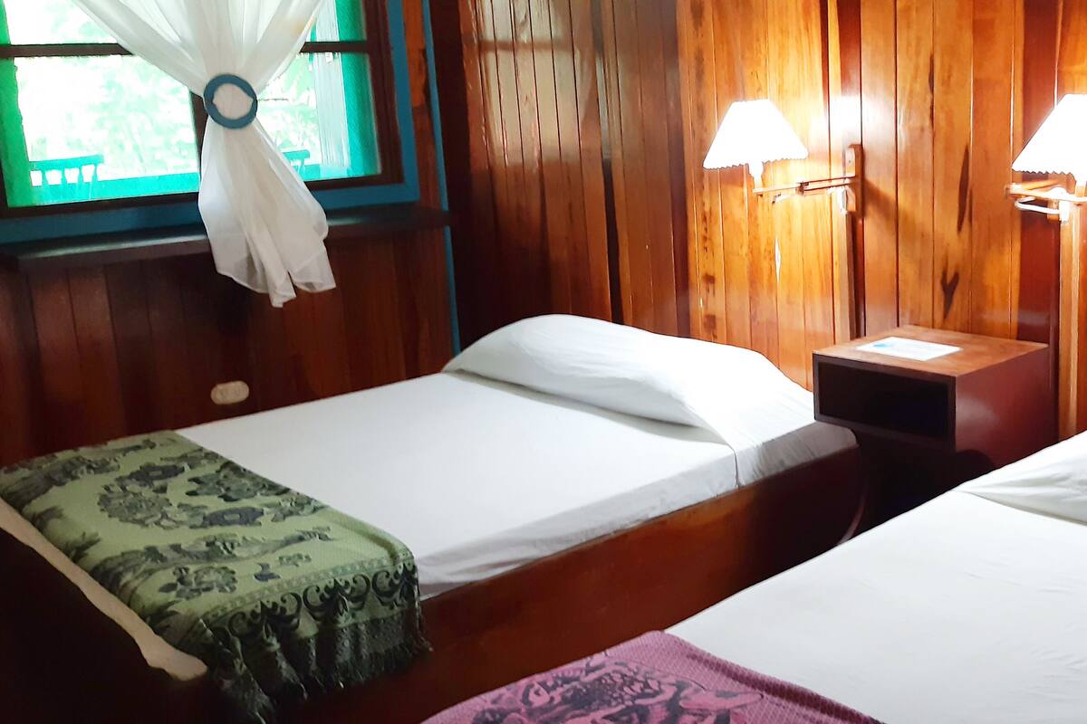 Una habitación con dos camas individuales, paredes de madera, una ventana con cortina y dos lámparas fijadas a la pared.