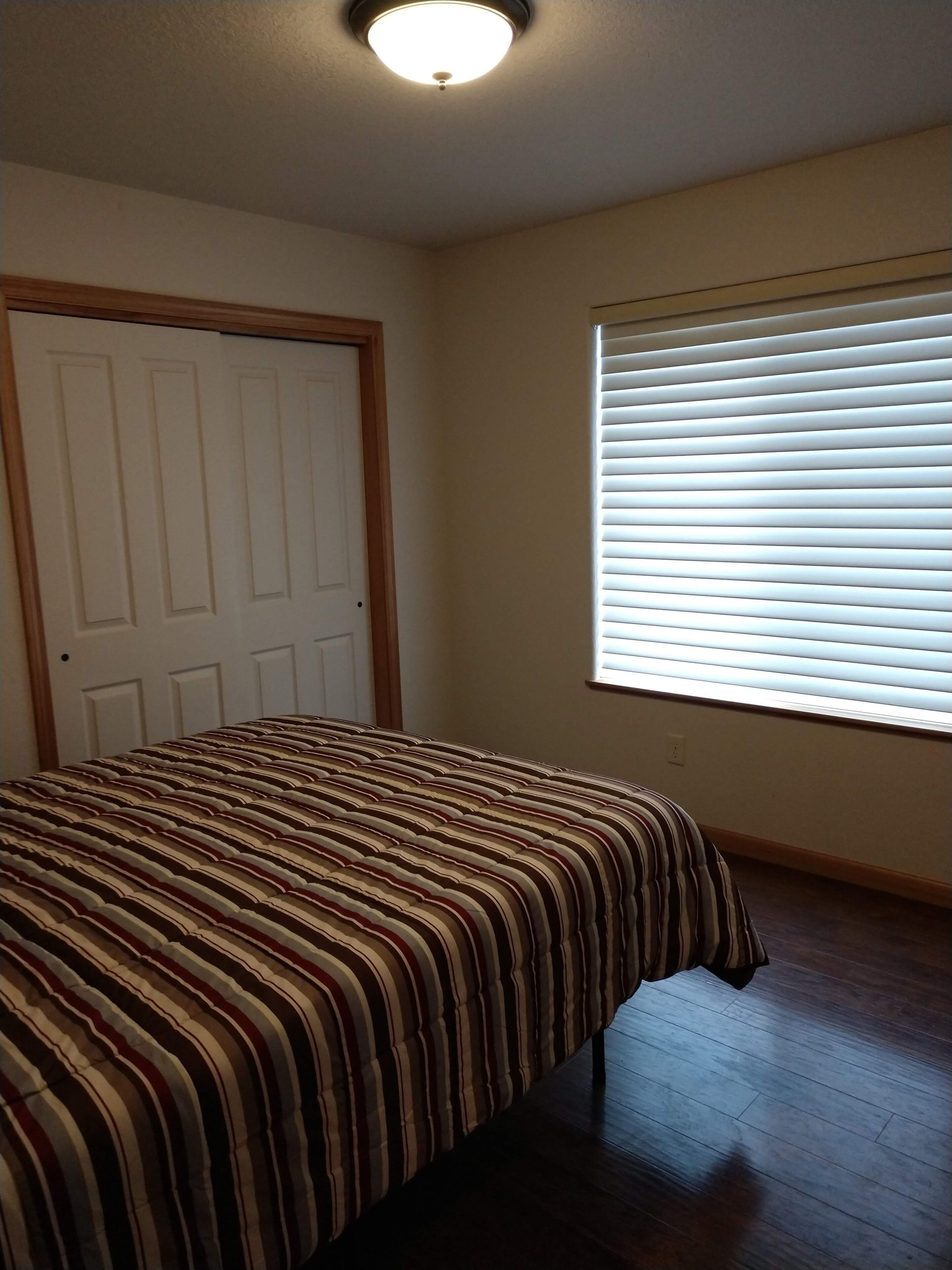 A bedroom with a bed, a door, a window with blinds, and a ceiling light.
