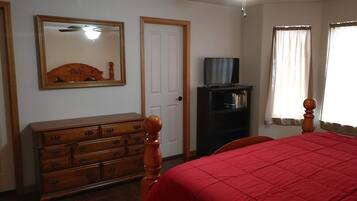 Een slaapkamer met een houten dressoir, een bed met een rode deken, een plafondventilator, een televisie op een standaard en een deur.