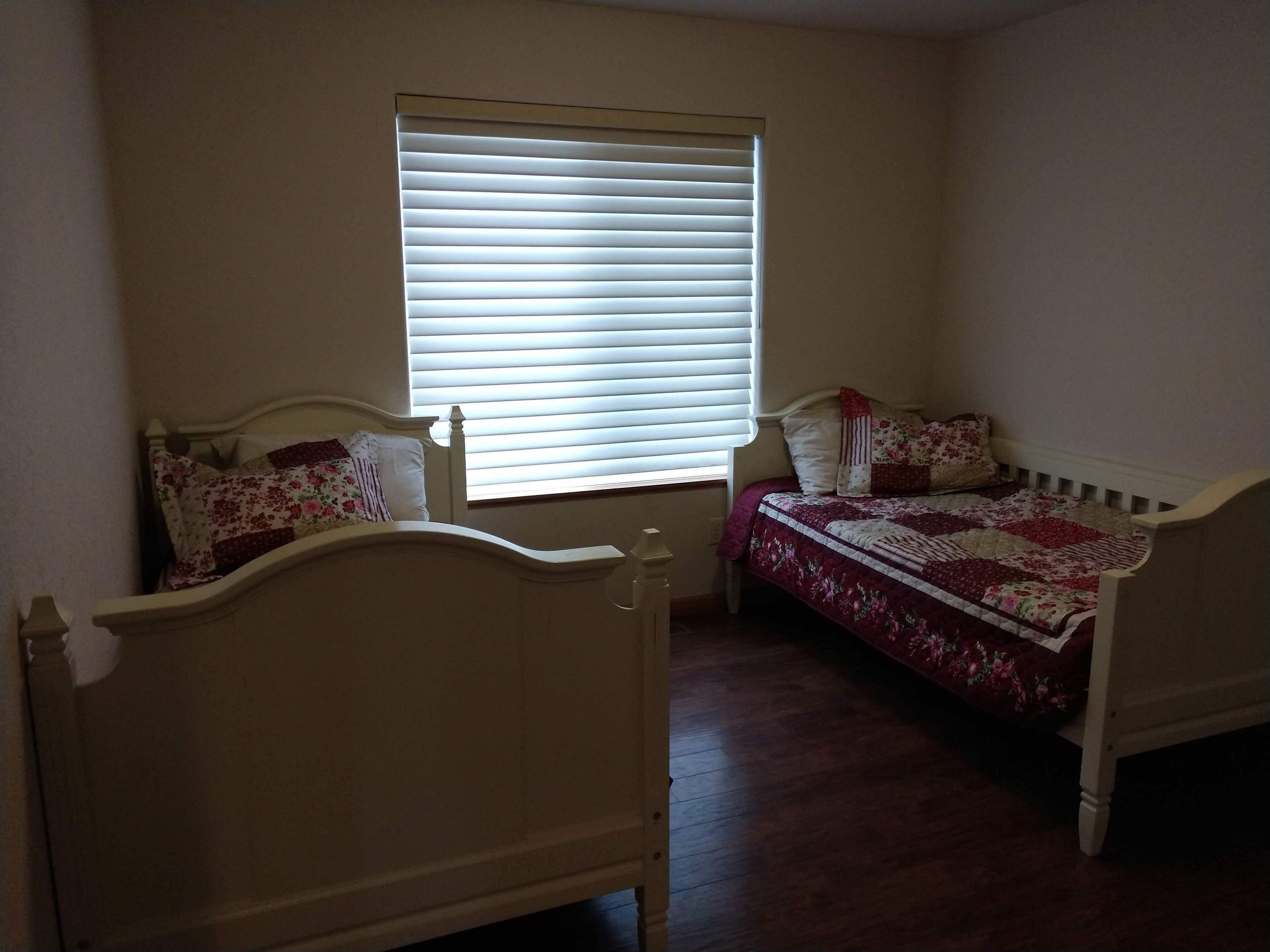 A small, well-lit room with two single beds, a window with blinds, and a radiator.