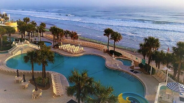 A resort pool with a curved design, surrounded by palm trees and lounge chairs, with a view of the ocean. - 1 Bd Condo, Wide Open Ocean F U N! Atlantic_1BD (Daytona Beach)