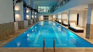 An indoor swimming pool with clear lanes, surrounded by walls with a stone-like texture, and a balcony overlooking the pool area. - The CLIFF - 4th-floor Luxury Condo on the Beach - Luxury Beachfront Resort  (Cupecoy)