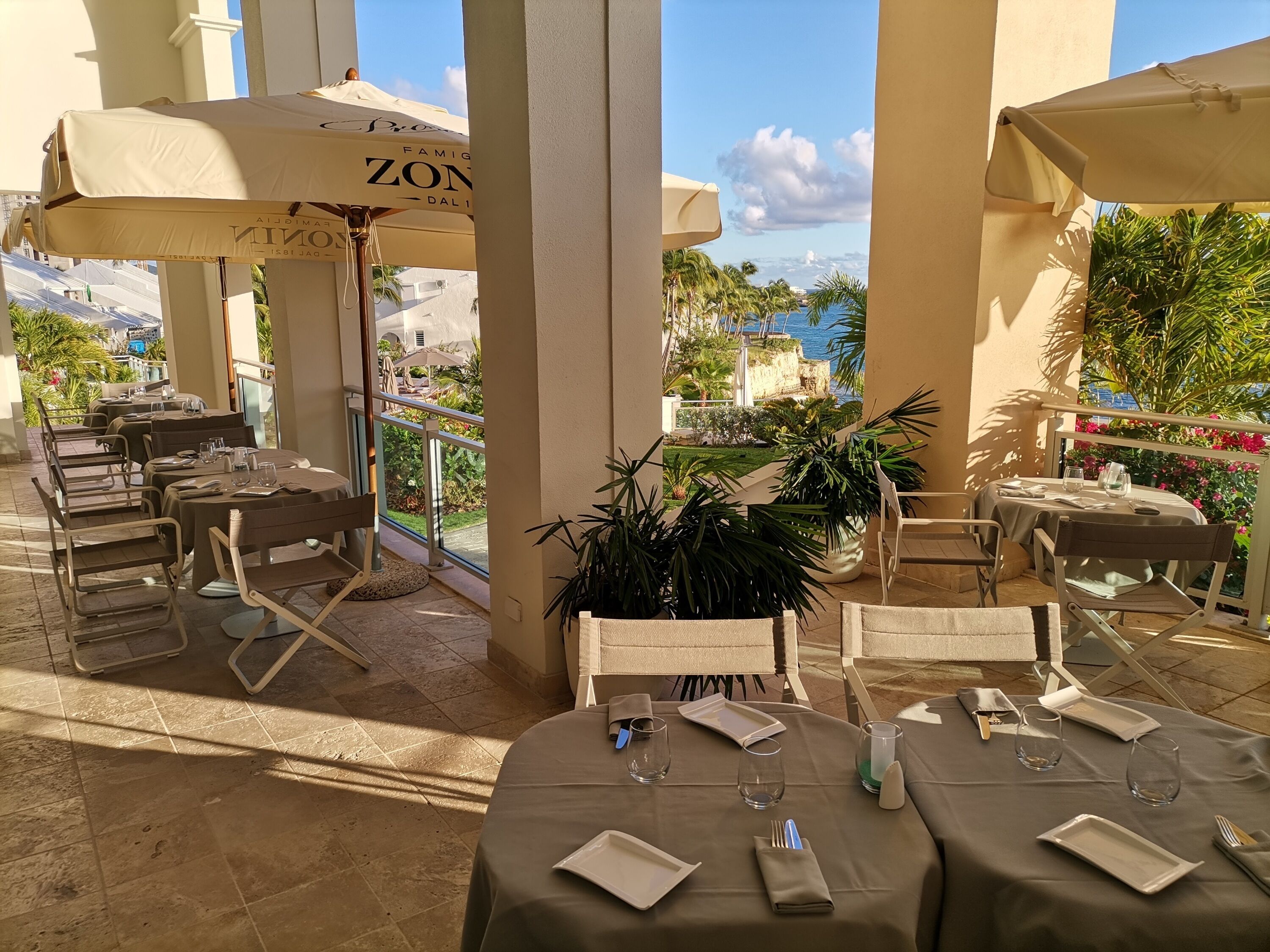 An outdoor dining area with tables and chairs, umbrellas, and a view of the sea.