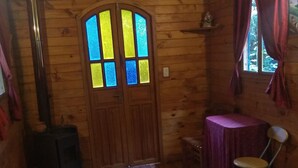 A wooden room with a bed, a table, a chair, a fireplace, and a window with curtains. - Hosteria La Roulotte Mindo (Mindo)
