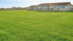 A large, open grassy field with a single-story building featuring a red umbrella and a fenced patio area. - 2 bedroom accommodation in Bacton (Bacton)