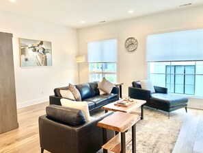 A modern living room with a black leather sofa, a wooden coffee table, and a blue armchair. There is a large window providing natural light and a clock on the wall.