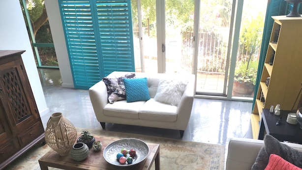 A living room with a sofa, a coffee table with decorative items, and a view through a glass door to an outdoor area with a blue gate. - Stylish Comfortable Central Artists Home (Glen Waverley)