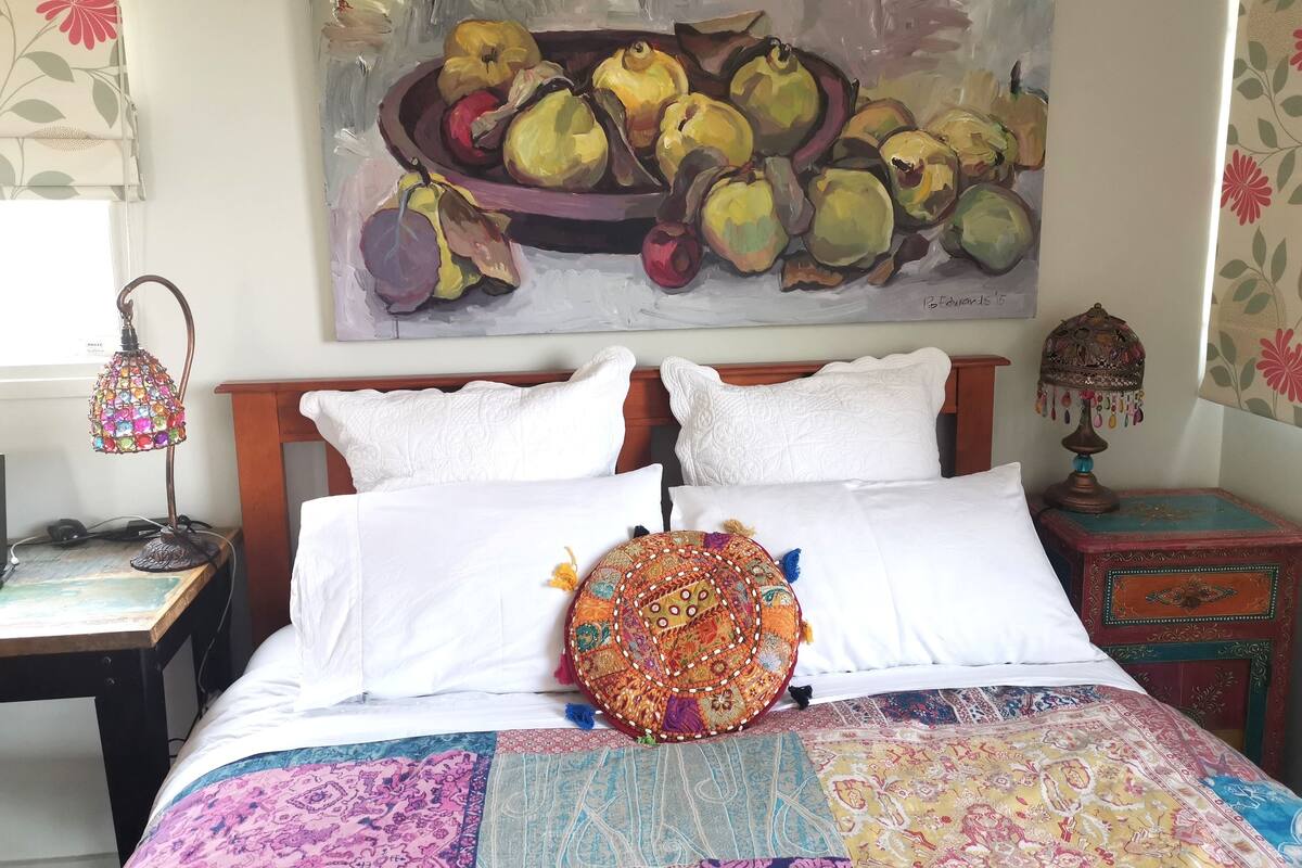 Quarto com cama, duas almofadas brancas, uma almofada colorida com bordado, colcha estampada, pintura de frutas na parede, abajur decorativo e mesa de cabeceira com luminária.