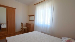 Un dormitorio con cama, armario de madera, silla, cómoda y ventana con cortinas traslúcidas.