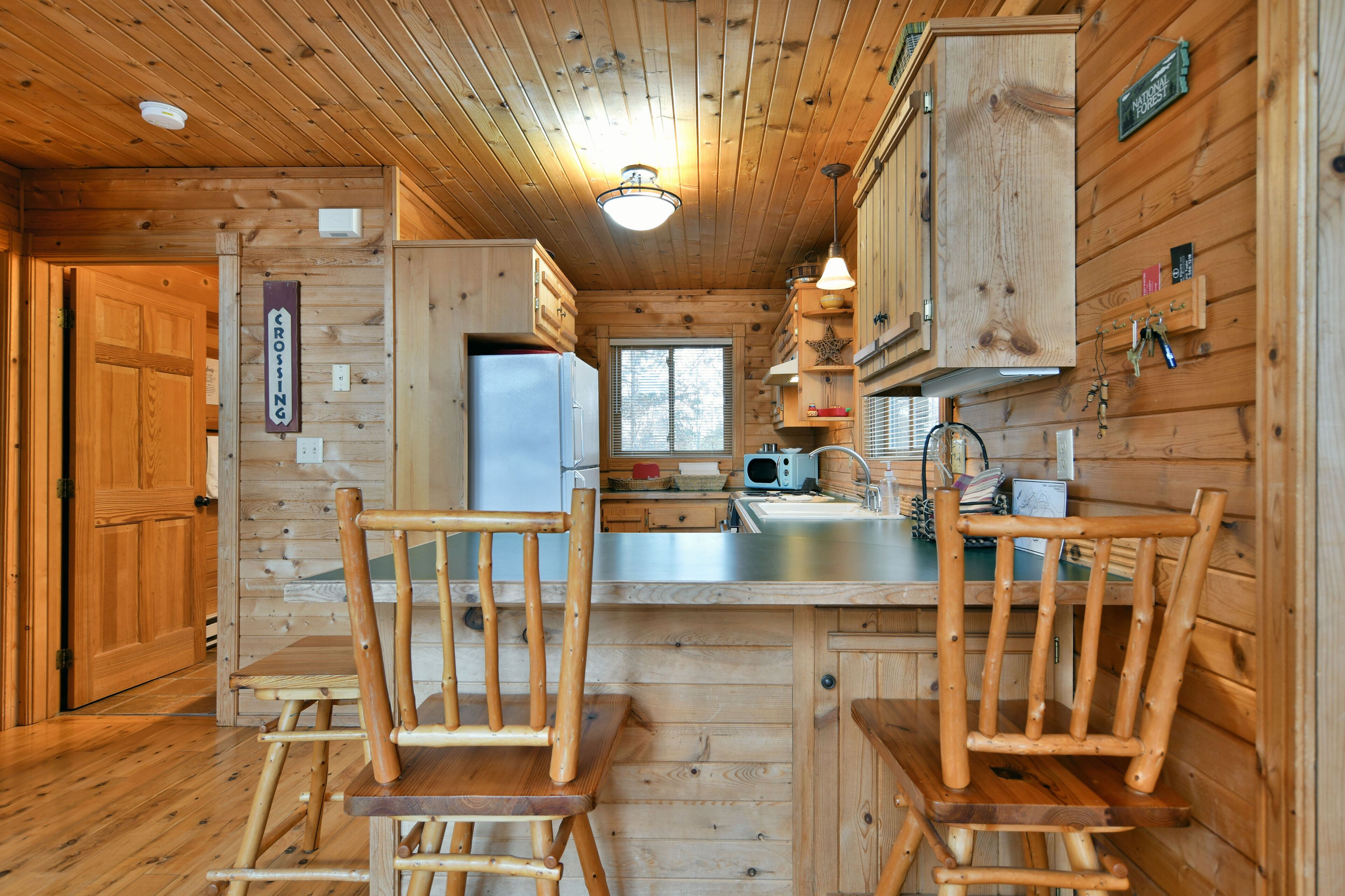 Interior de una cabaña de madera con zona de cocina, sillas de madera y un refrigerador.
