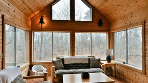 Interior acogedor de una cabaña con cama en altillo, un sofá, una mesa y grandes ventanales que dan a un bosque.