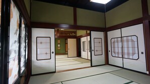 Une pièce de style japonais traditionnel, avec un sol recouvert de tatamis, des portes coulissantes ornées de motifs décoratifs et une vue sur une autre pièce dont le plafond est en bois.