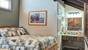 Ein Schlafzimmer mit einem hölzernen Bett, einer gesteppten Tagesdecke, einem gerahmten Bild an der Wand und einem Badezimmer mit einem Holzwaschtisch und einem Spiegel.