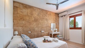 A bedroom with a stone wall, a bed with white bedding, a bedside table with a lamp, a small desk with a mirror, and a window with curtains. - Rural retreat in the heart of nature: enjoy tranquility and privacy (Pollença)