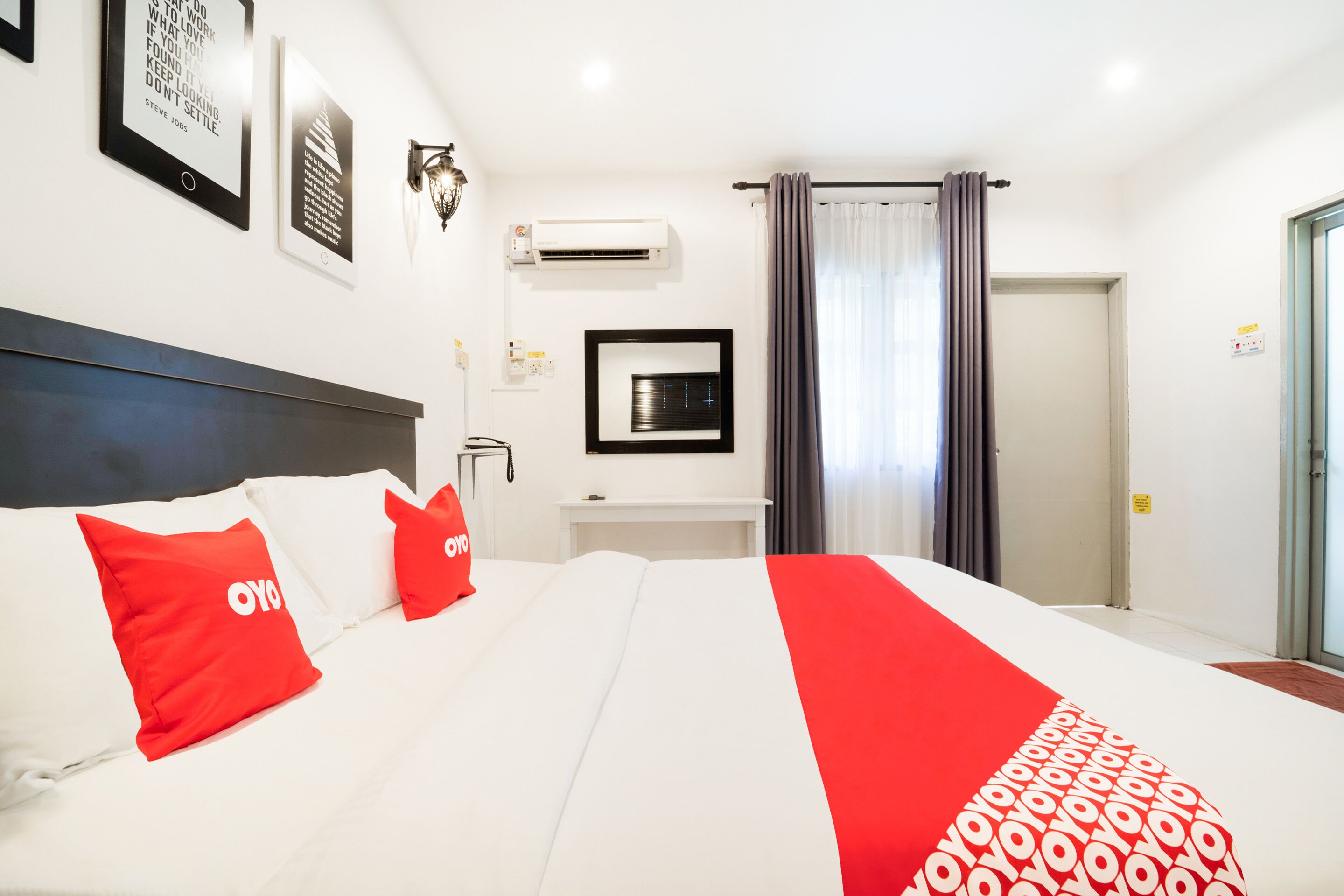 A hotel room with a large bed, red and white bedding, a mounted TV, and a window with curtains.