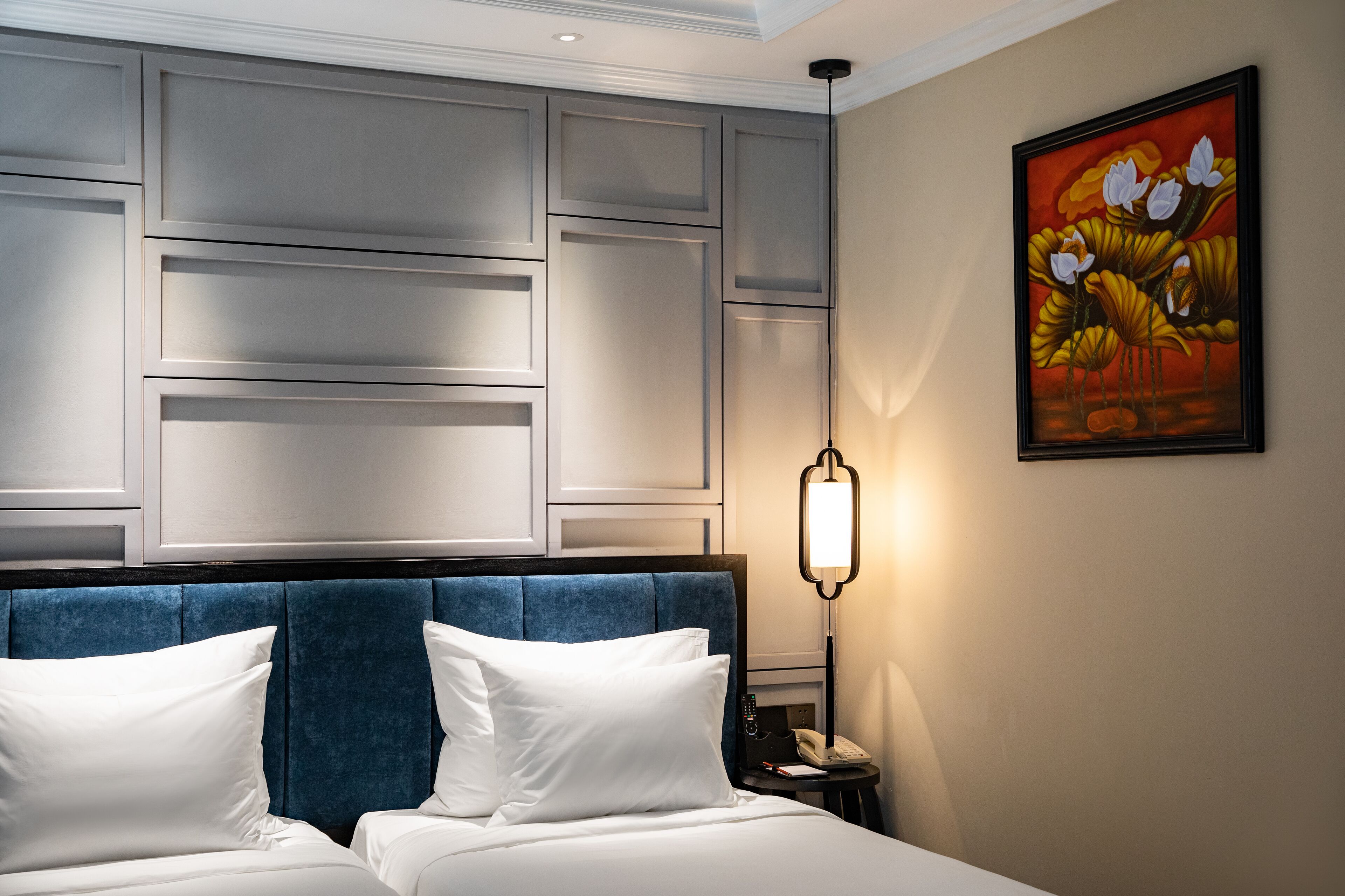 A hotel room with a large bed, a headboard with a blue upholstered panel, a bedside table with a lamp, and a painting on the wall.