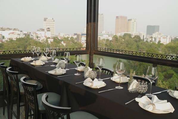 A dining table set with glasses, plates, and cutlery, with a cityscape view through large windows. - Solaria Hotel (Hanoi)