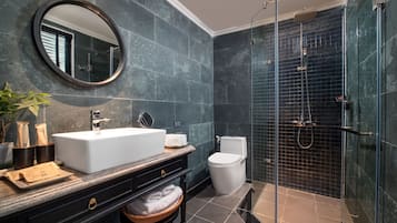 A modern bathroom with a large white sink, dark marble walls, a glass-enclosed shower, and a round mirror.