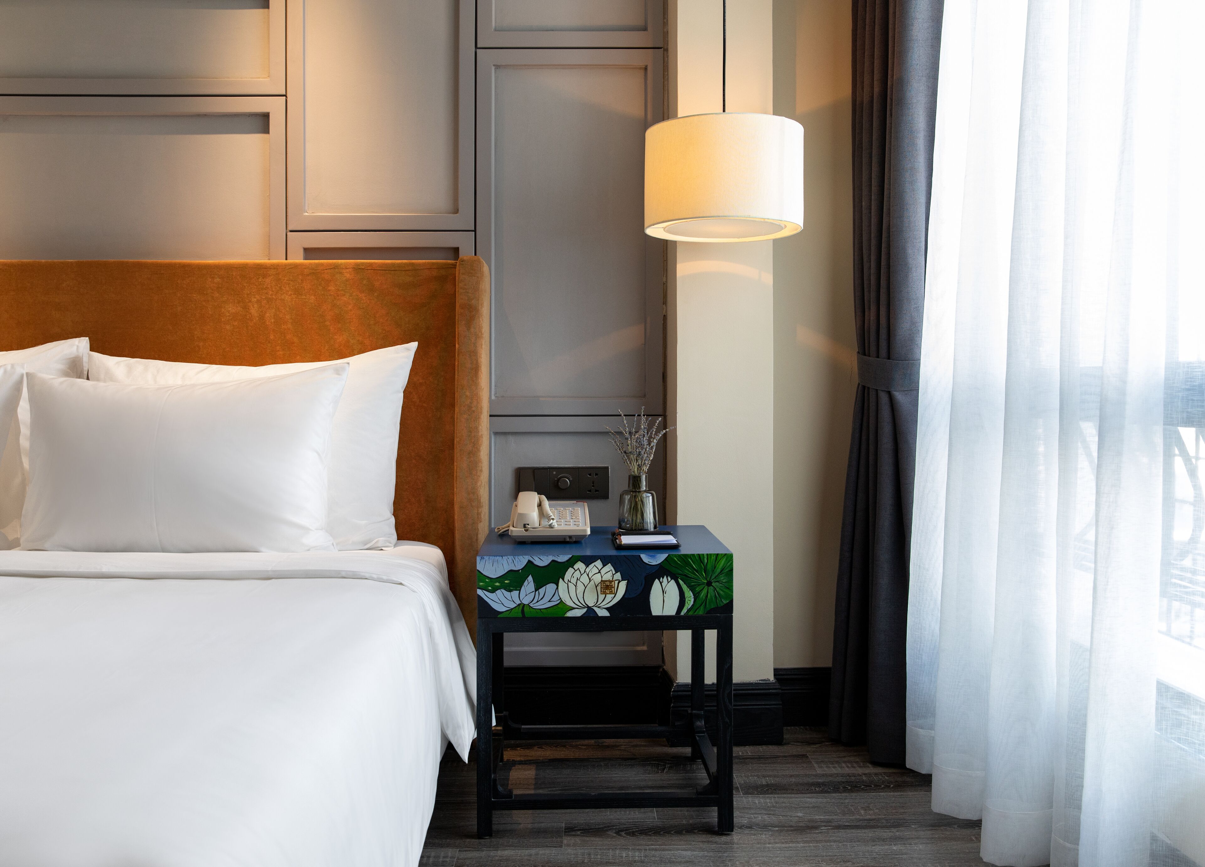 A hotel room with a bed, a bedside table with a lamp, a phone, and a vase with dried flowers.
