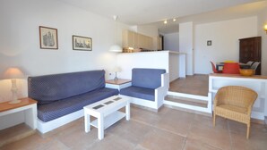 Ein Wohnzimmer mit Sofa, Sessel und kleinem Tisch. Im Hintergrund ist eine Küchenzeile erkennbar.